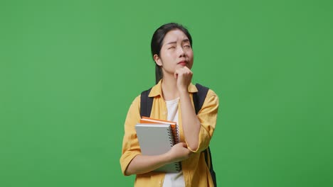 asian woman student with a backpack and some books thinking and looking around then raising her index finger while standing in the green screen background studio