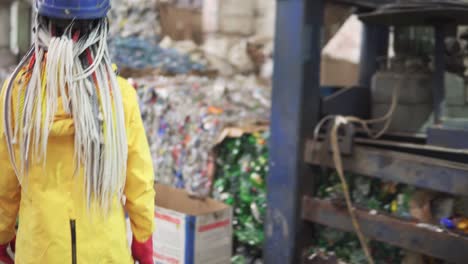 Woman-worker-in-yellow-and-transparent-protecting-glasses,-hard-hat-and-mask-working-with-equipment-on-recycle-plant.-Press-machine-with-used-plastic-bottles-and-differen-plastic-garbage.-Footage-of-automized-process