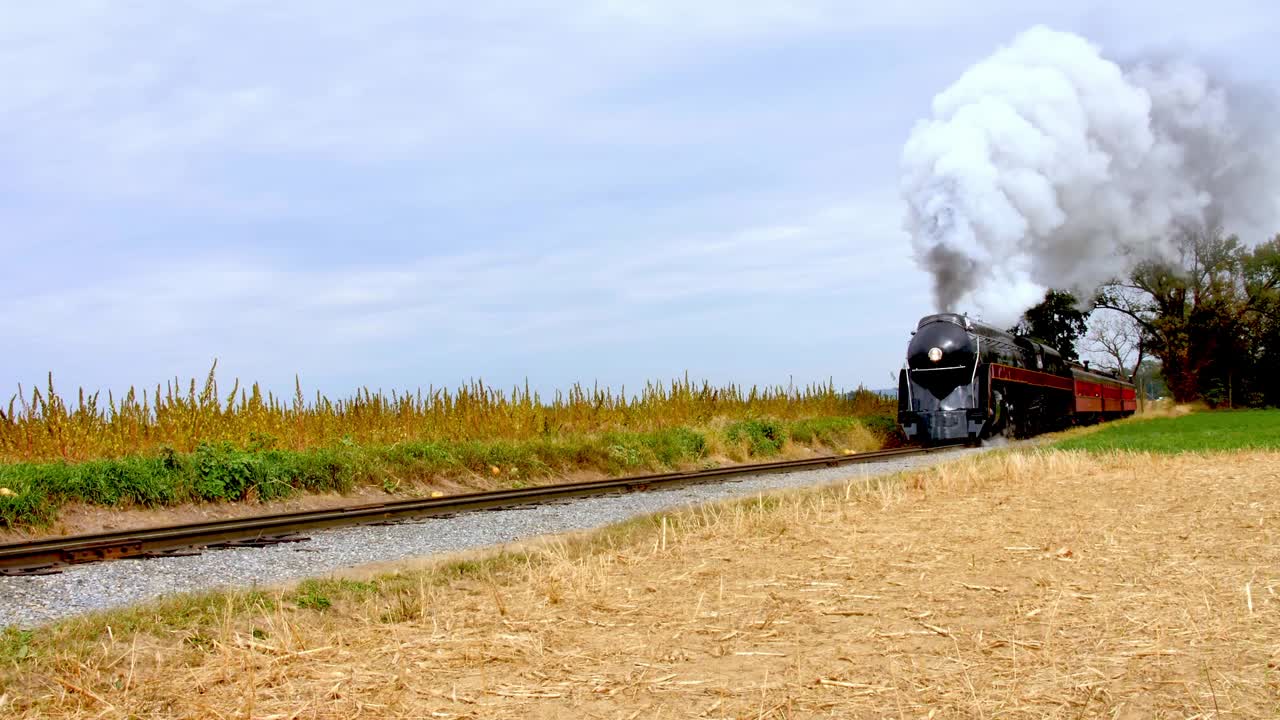 A vintage stream lined steam locomotive chugs along railway tracks surrounded by golden fields. The sky is partly cloudy, enhancing the picturesque autumn landscape.