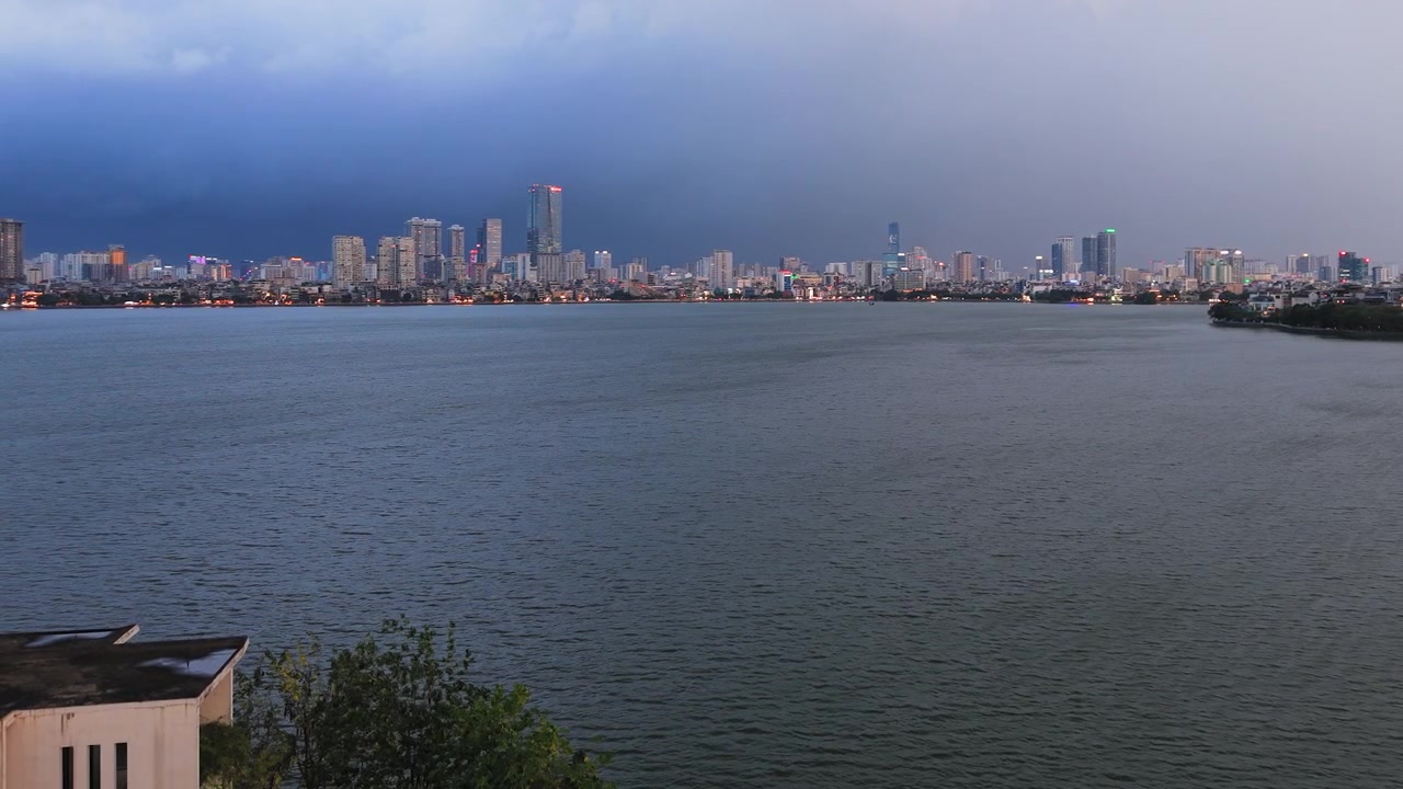 Un'ampia inquadratura drammatica di un temporale con fulmini che si avvicina allo skyline della città di Hanoi attraverso il Lago dell'Ovest, con nuvole scure e luci urbane