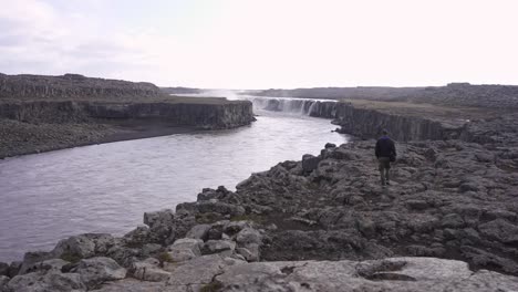 Viajero-Caminando-Por-La-Orilla-Rocosa-Del-Río-Hacia-La-Cascada