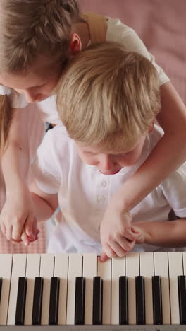 blonde little girl presses piano keys with crying brother fingers in bedroom upper view. toddler boy fatigued by difficult music lesson with sister