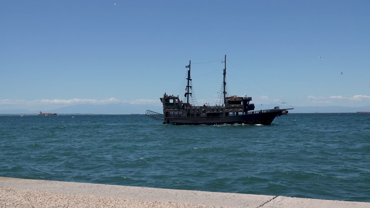 A vintage-style black pirate ship sails across the calm blue sea under a clear sky, with distant ships and mountain ranges visible on the horizon, evoking a sense of maritime adventure