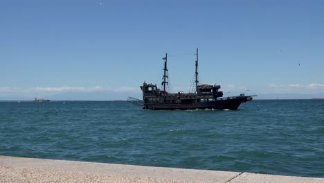 a vintage-style black pirate ship sails across the calm blue sea under a clear sky, with distant ships and mountain ranges visible on the horizon, evoking a sense of maritime adventure