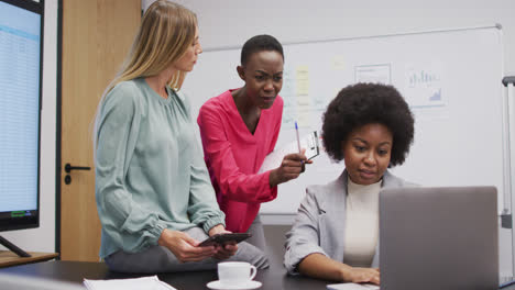 Tres-Empresarias-Diversas-En-Discusión-Usando-Una-Computadora-Portátil-Y-Una-Tableta-En-Una-Oficina