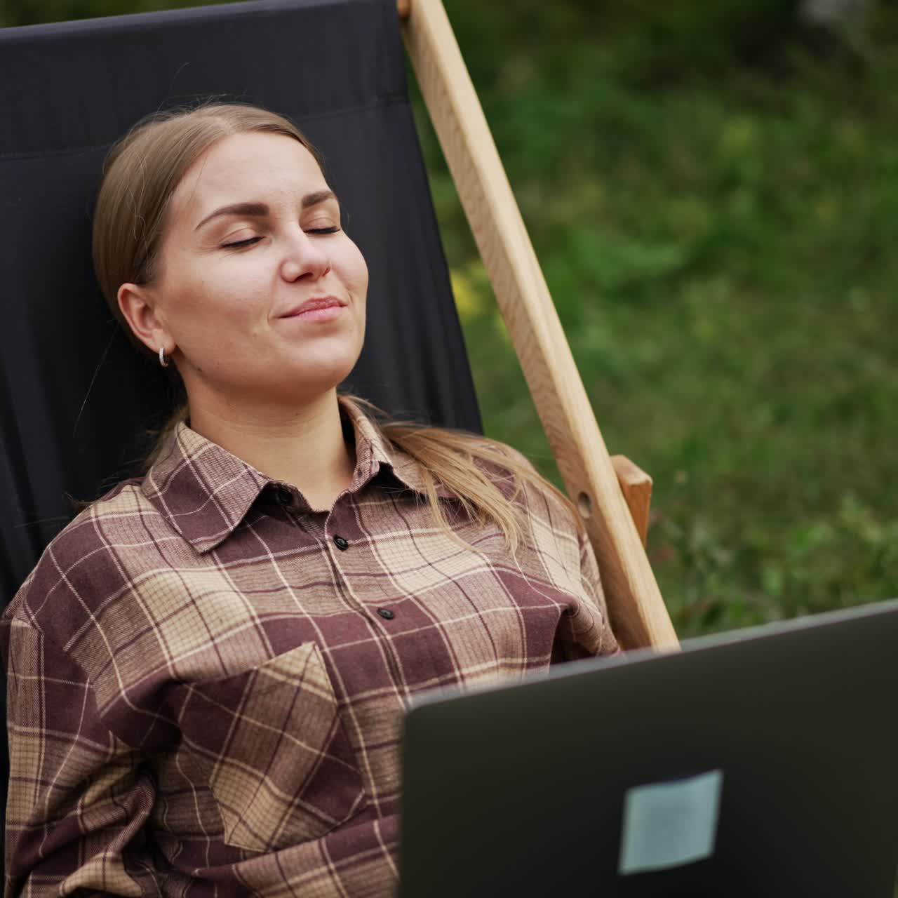 Positive smiling young woman sits in garden chair working on laptop. Lady distracts for a moment, closes her eyes to have a little break from remote work