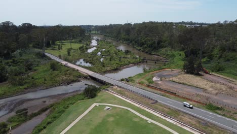 Dron-Descendiendo-A-Un-Parque-Verde-Que-Muestra-Un-Pequeño-Río-Y-Un-Puente-Con-Autos-Pasando