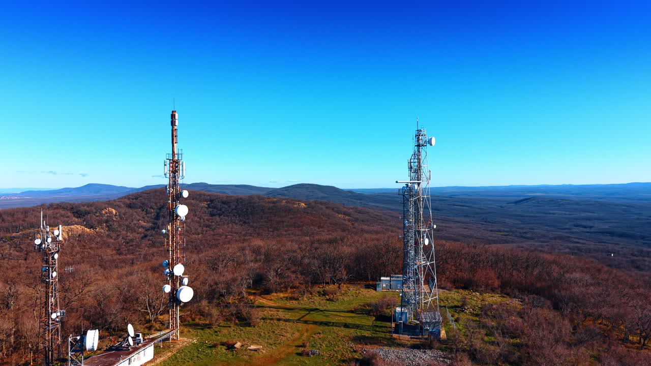 Telecommunication towers on forested hill under clear blue sky. Group of cell phone and radio masts atop hill with dry autumn trees and horizon view