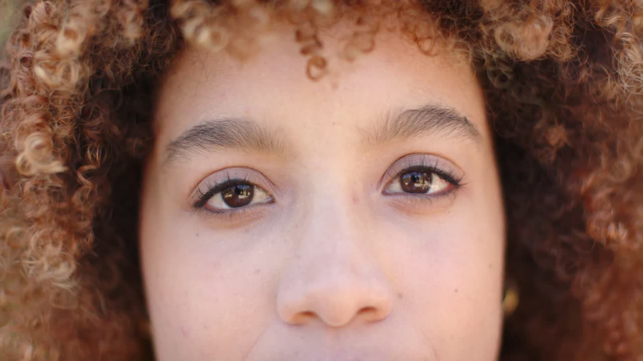 retrato de una mujer biracial feliz con el cabello rizado mirando a la cámara en el jardín en cámara lenta.