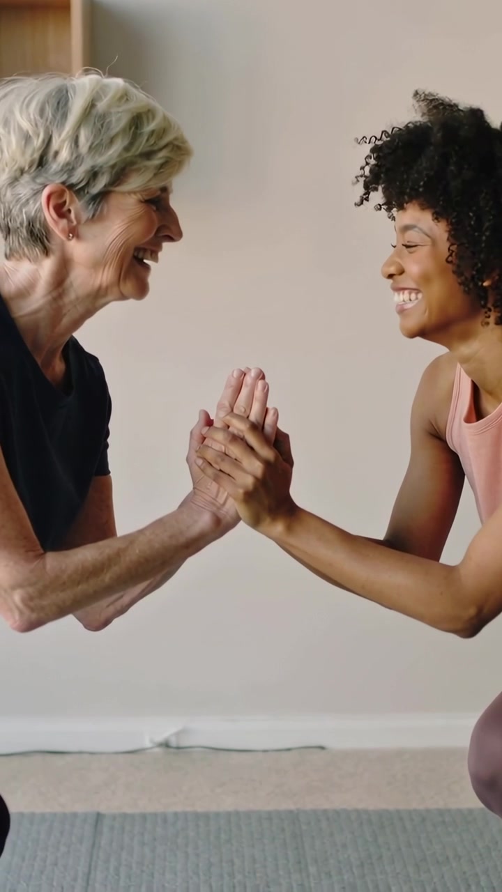 Vertical video: Pressing palms two women starting low squat on mat for balance, wearing pink top