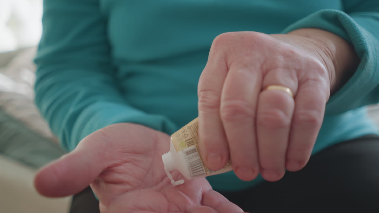 Old woman in green applying ointment to her hand while sitting on bed. Soft light streams in from the window, creating a calm and serene atmosphere. A moment of self-care in a peaceful space