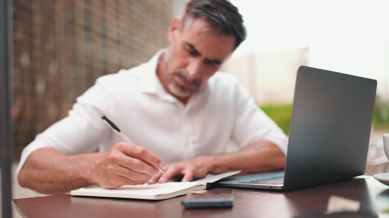 Man working on laptop and writing in notebook at outdoor cafe