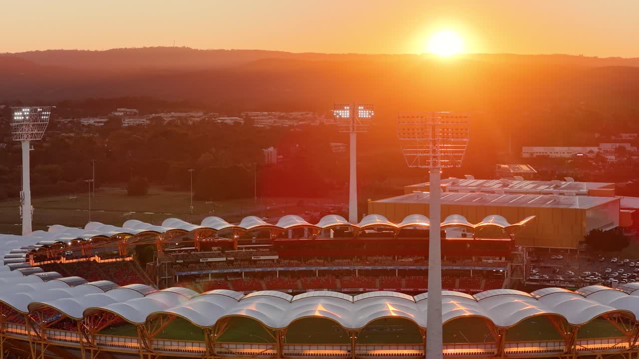 Drone footage glides over a contemporary football stadium at sunset, capturing stadium lights, stands, and nearby river in warm golden hour lighting