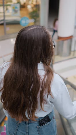 바쁜 쇼핑몰에서 레일에 손을 고 내려가는 에스컬레이터에서 머리카락을 과시하는 젊은 성인의 뒷면, 공공 쇼핑 공간에서 움직임과 캐주얼 우아함을 포착합니다.