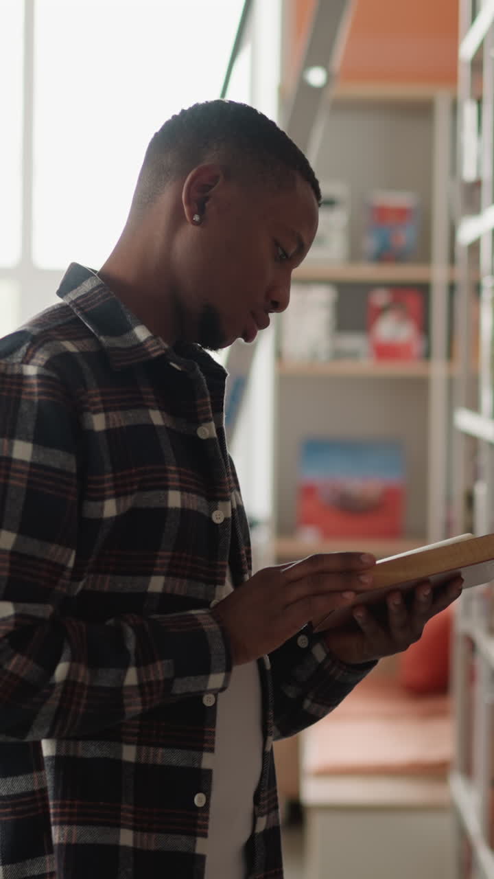 lector masculino pasa por un libro de pie al lado de una estantería. joven investigador afroamericano da la vuelta a las páginas buscando información para un proyecto científico. biblioteca universitaria