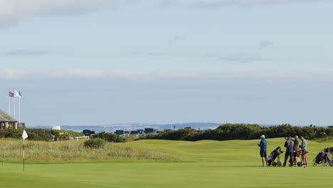 a group of golfers with equipment walk across a sunlit fairway toward a traditional stone clubhouse under partly cloudy skies, captured in a smooth lateral pan