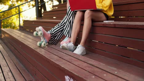 close-up shot: a girl in striped pants and roller skates and a girl in shorts and white sneakers sit on a brown bench and work on a red laptop in a park in summer