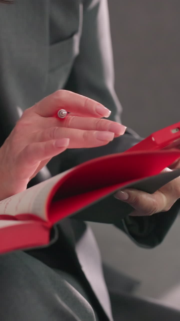 Businesswoman writing in red notebook, holding pen, modern office environment, gray suit, focused on task, productivity, planning, decision-making, workspace, corporate setting, concentration