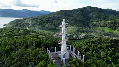 Breathtaking-Aerial-View-of-White-Lighthouse-atop-mountain-in-lush-rainforest-with-vast-sea-and-clouds-in-background