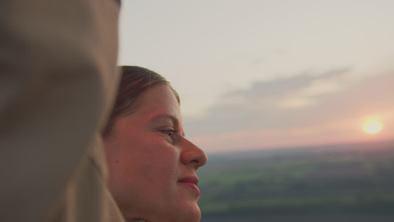 Close up of woman face in warm morning light, partially framed by arm as she increases metal burner inside hot air balloon, with soft sky and horizon fading in background