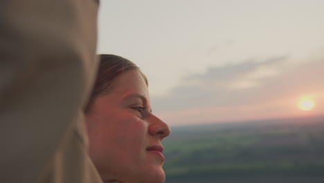 close up of woman face in warm morning light, partially framed by arm as she increases metal burner inside hot air balloon, with soft sky and horizon fading in background