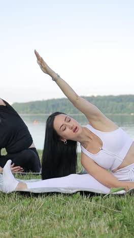 two beautiful slender young women are doing yoga she is sitting on a mat and raising her arms up two beautiful slender young women are doing yoga she is sitting on a mat and raising her hands up in the city park women practicing yoga outdoors on a sunny day
