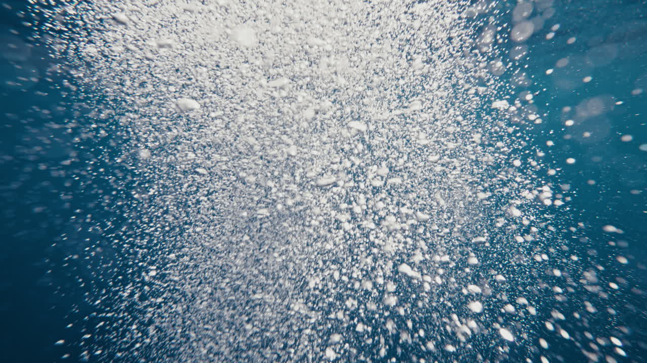 Pyöriviä ilmakuplia merenpinnalla, makrokuvassa yksityiskohdat nousevat pintaan valonsäteiden valaisemassa tekstuuripylväässä