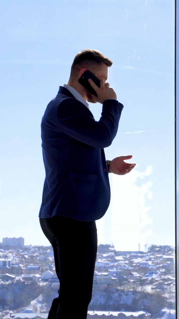 Businessman having a conversation through the phone. Young man in elegant suit standing near the large office window and talking the phone. Rear view. Vertical video