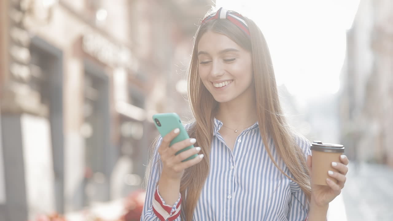 Giovane donna sorridente che beve un caffè mentre usa lo smartphone per strada