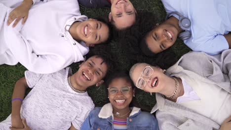 relajados adolescentes amigos multirraciales acostados juntos en la hierba verde con los ojos cerrados, despertarse y sonreír mirando a la cámara