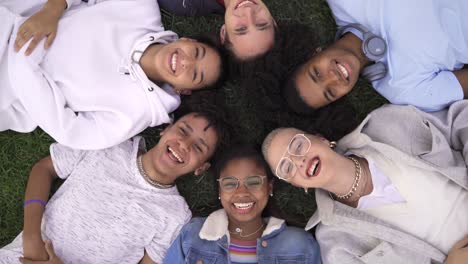 relaxed teenager multiracial friends lying together on green grass with eyes closed, wake up and smile looking to camera