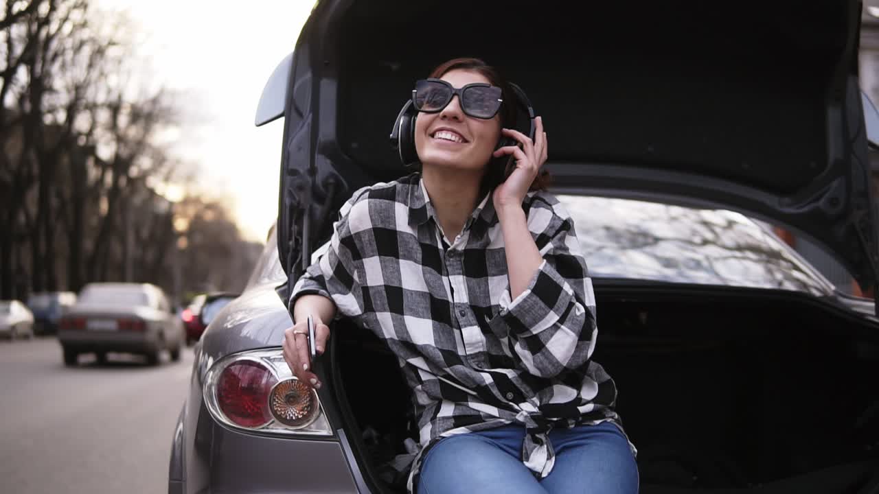 Smiling brunette listen to the music in headphone while sitting in the open car's trunk among the road. Cell phone. Good mood