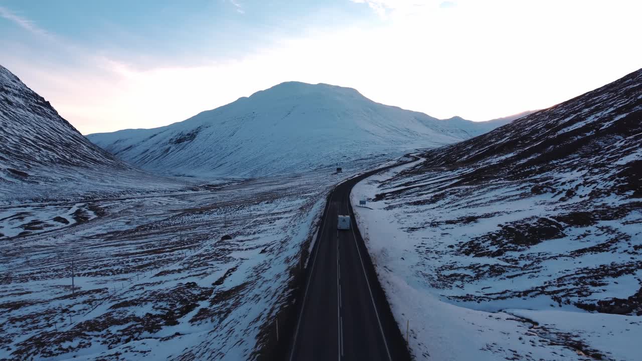 An aerial shot captures a motorhome driving along a winding road that cuts through a majestic, snow-covered mountain valley in Iceland