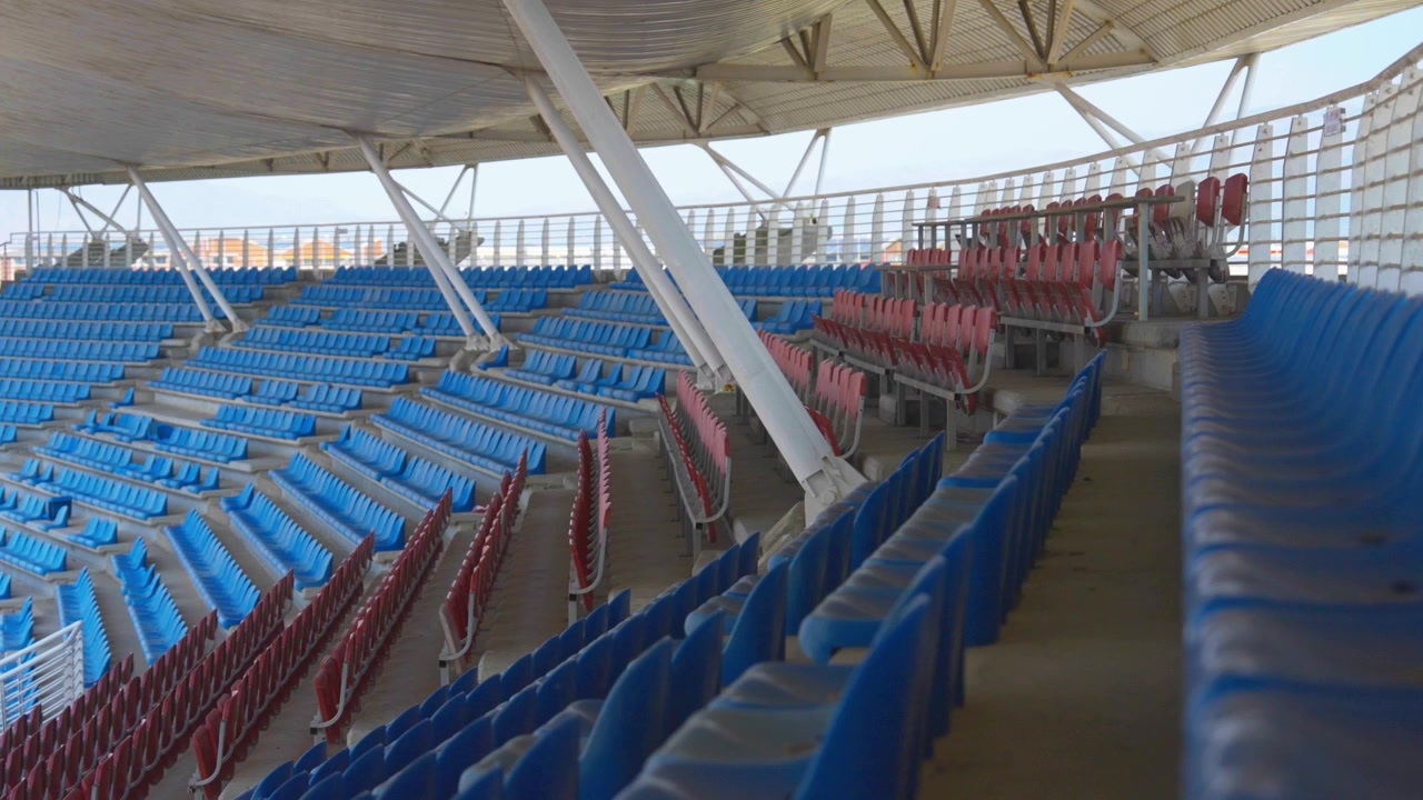 A wide shot captures rows of empty blue and maroon plastic seating curving beneath a corrugated white stadium roof, supported by angled steel beams.