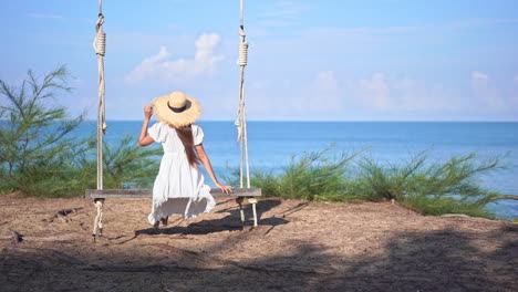 Auf-Einer-Riesigen-Holzschaukel-Sitzend-Schaukelt-Eine-Junge-Frau-Mit-Dem-Rücken-Zur-Kamera-Hin-Und-Her,-Während-Sie-Auf-Das-Meer-Blickt