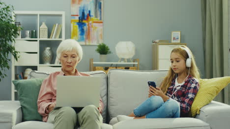 anciana sentada en el sofá y trabajando en la computadora portátil mientras su nieta se sienta al lado y escucha la música, luego la niña se los quita y habla con la abuela