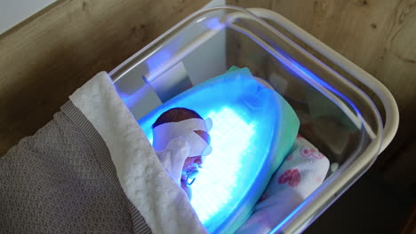 high bilirubin treatment for a newborn child. tiny baby lies on a ultraviolet lamp in a crib covered with blanket. top view.