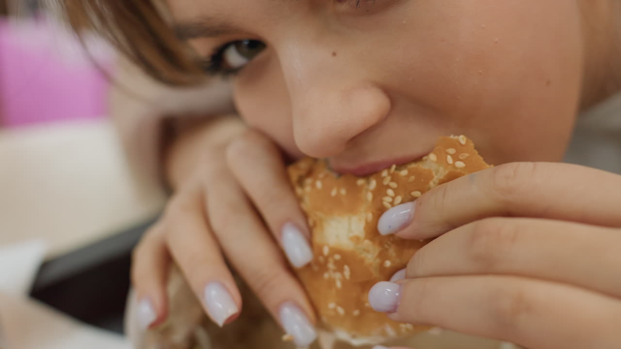 Primer plano de una chica caucásica comiendo una hamburguesa con actitud divertida, mirando directamente a la cámara, detalle del pan con sésamo, esmalte de uñas en los dedos, momento íntimo de tentempié, expresión pícara, migas de comida visibles