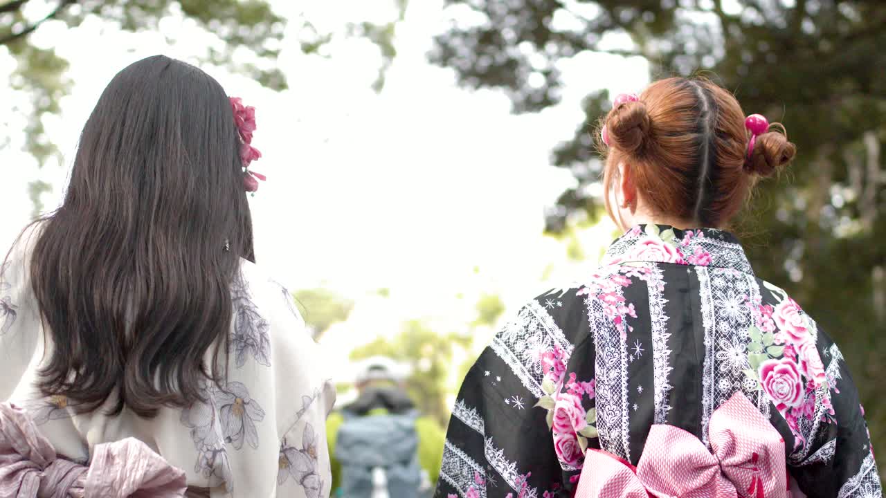 Hai người phụ nữ mặc yukata truyền thống họa tiết hoa đi dạo trong một công viên sáng sủa với những hàng cây xanh tươi tốt
