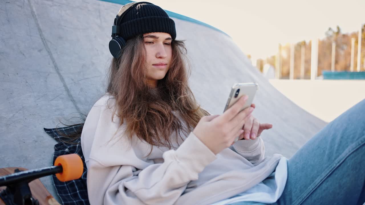retrato de cerca de una hermosa patinadora en un sombrero de punto y auriculares escuchando música. la niña está descansando sosteniendo un teléfono inteligente en las manos, su patineta se encuentra cerca