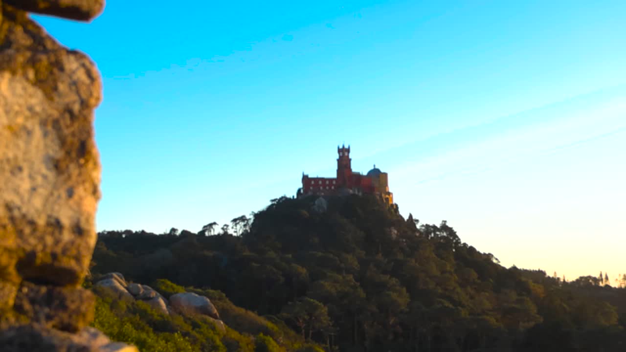 Cinematic sunset video filmed from the Moorish Castle in Sintra, detailed historic stone walls in the front with Pena Palace visible on a distant hill and mountain landscape under warm evening light