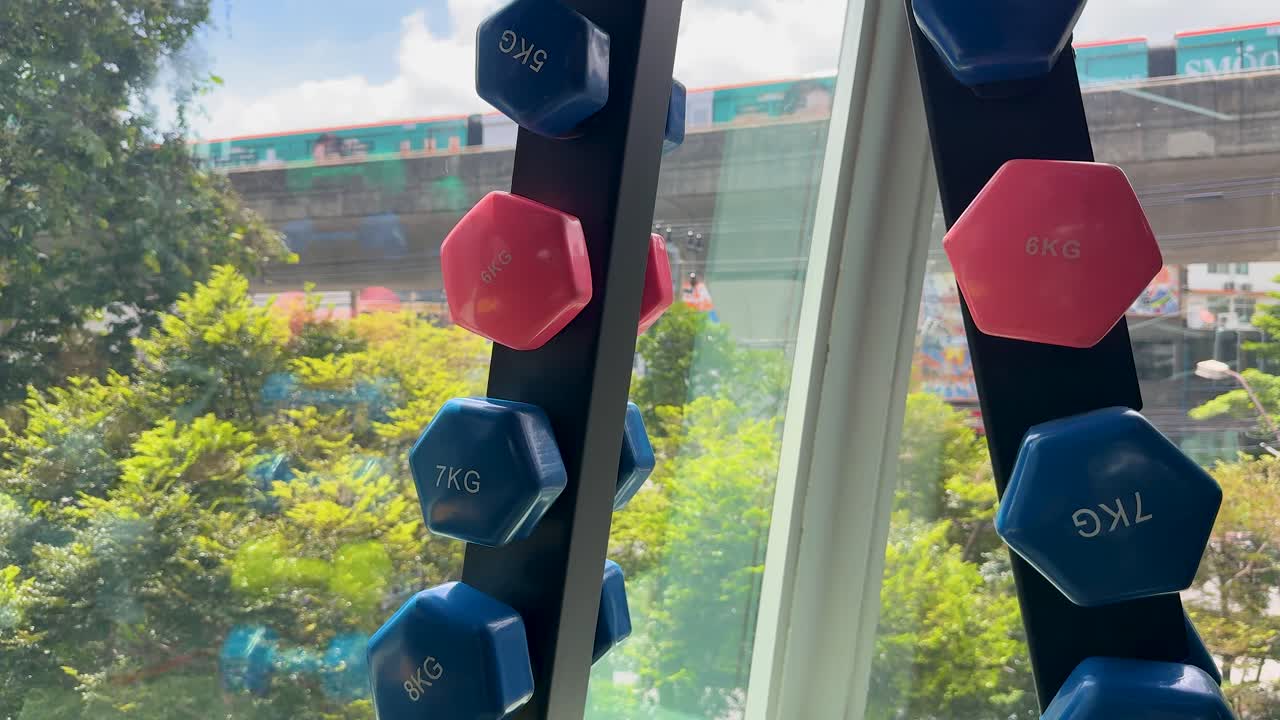 A camera smoothly circles a colorful dumbbell rack near a large window in a bright gym, revealing city and greenery outside
