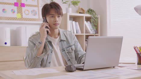 asian designer man uses his smartphone to communicate. work on projects and papers. and work from home on a desk with a laptop.