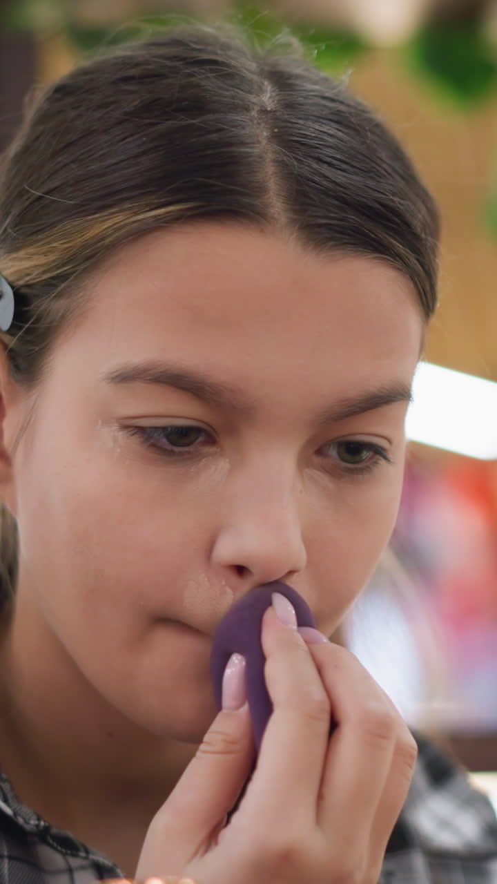 Adolescente aplicándose base de maquillaje con una esponja morada, iluminación interior suave, camisa de cuadros, expresión concentrada, difuminando desde la barbilla hasta las mejillas, primer plano con espejo, preparación informal en casa para ir al colegio