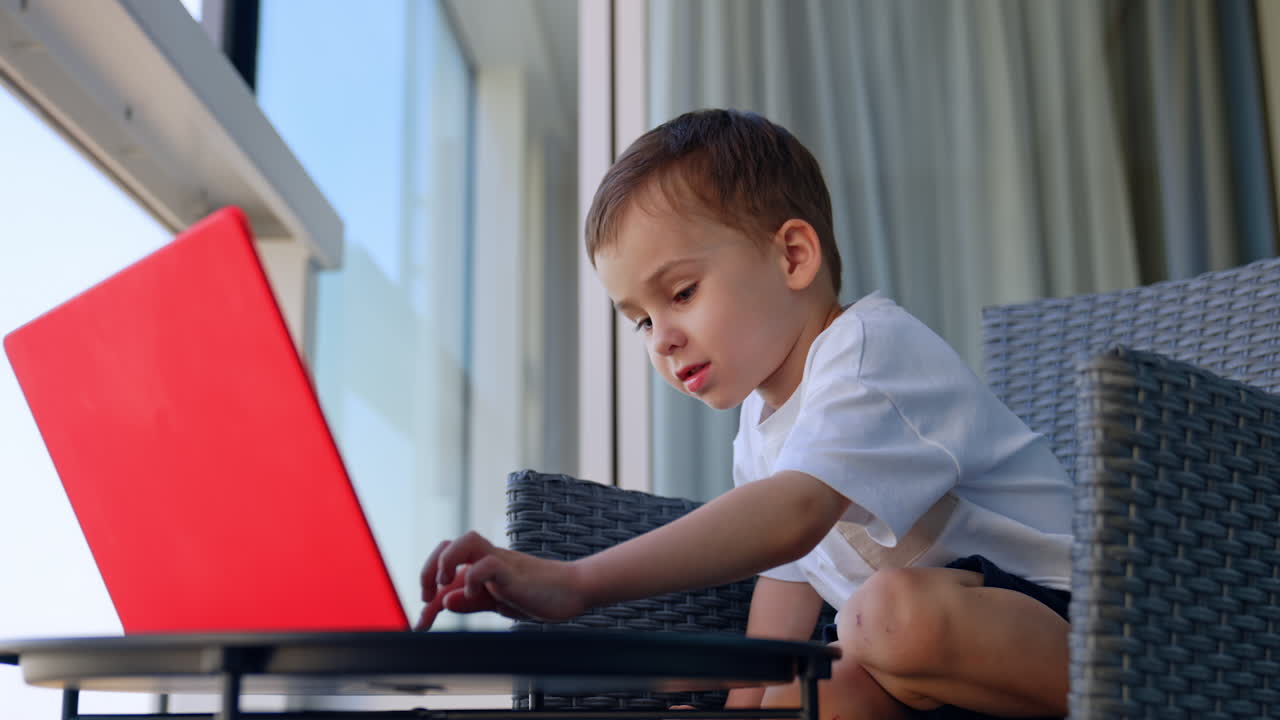 Boy sitting on chair using laptop on balcony. Little boy in white t-shirt using laptop on balcony with panoramic city view in background