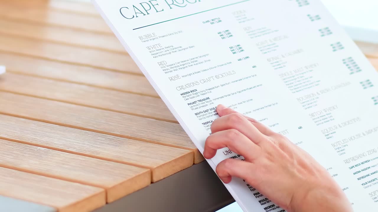 Close-up of hands flipping through a drink menu on a wooden table in bright lighting.