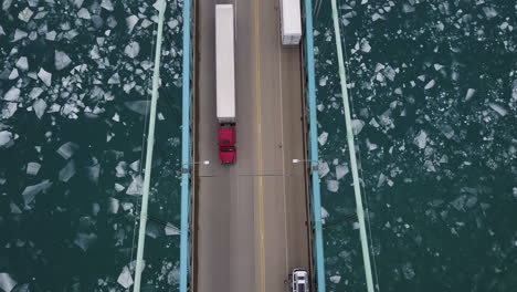 Aerial-top-down-shot-of-transport-trucks-crossing-a-bridge-over-icy-blue-water