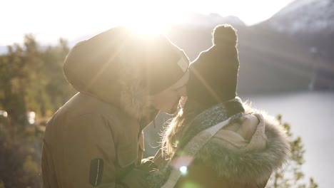 young couple standing embrace kissing, looking landscape sunny winter day. woman and man in warm clothes standing in front beautiful landscape of norwegian mountains peaks and winter lake. lens flares ona background