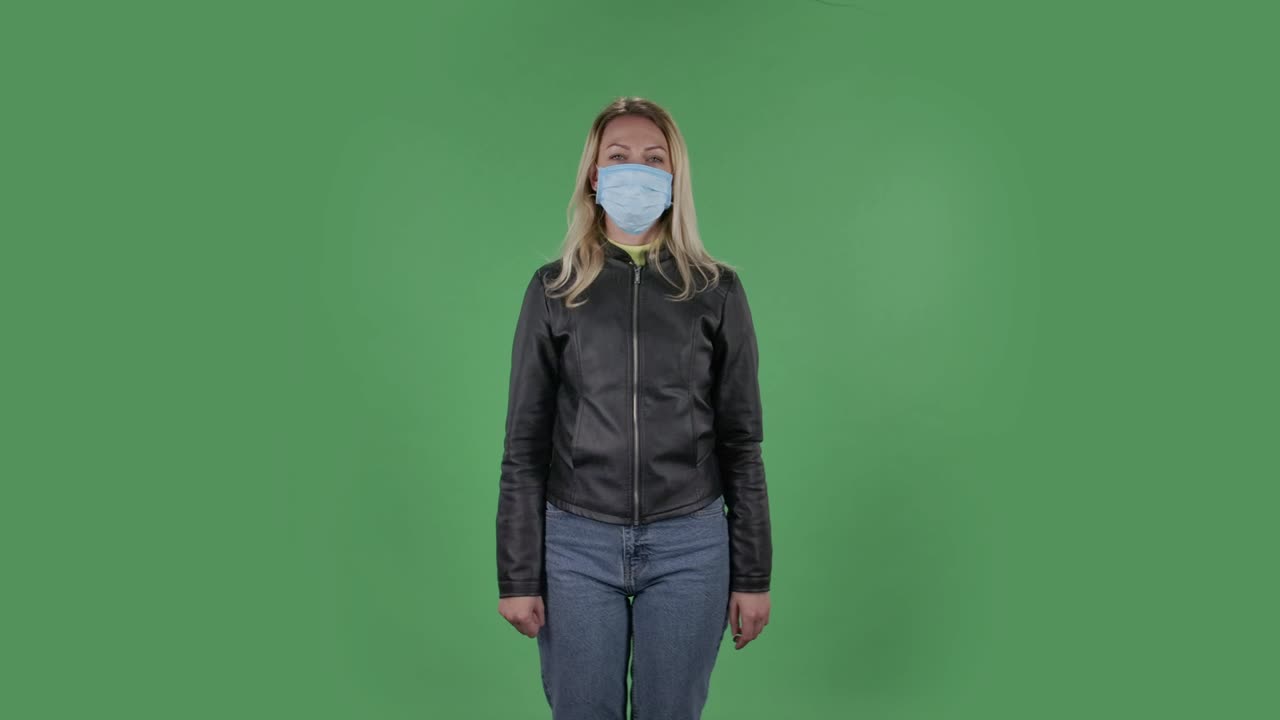 의료 보호 얼굴 마스크를 입은 아름다운 젊은 여성의 초상화는 카메라를보고 주먹을 보여 주고, 한, 두, 세, 네, 다섯 손가락을 세고, 검은색 재과 청바지를 입은 느슨한 머리카락을 가진 금발은 스튜디오에서 녹색 화면에 건강 보호.