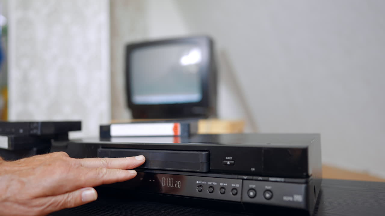 Male hand inserts VHS cassette into the player. Man presses the button and screen switches on at backdrop